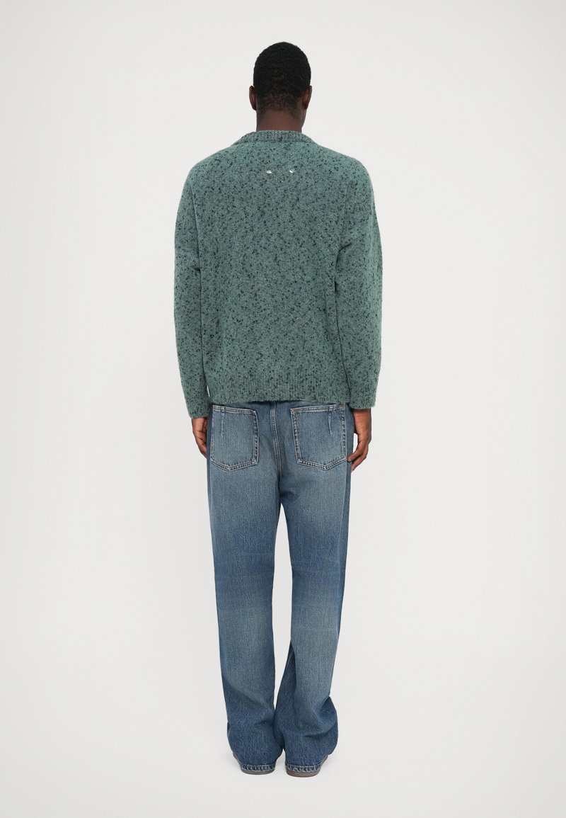 Green textured sweater with long sleeves, crew neck, and drop shoulders, paired with light blue, loose-fitting jeans with back pockets.