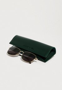 Estuche para gafas de sol Lacoste verde oscuro parcialmente abierto con gafas de sol de montura dorada y lentes tintados oscuros descansando a su lado sobre una superficie blanca.