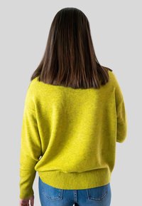 Persoon met steil bruin haar in een felgele trui en blauwe jeans, met de rug naar de camera gekeerd tegen een effen grijze achtergrond.