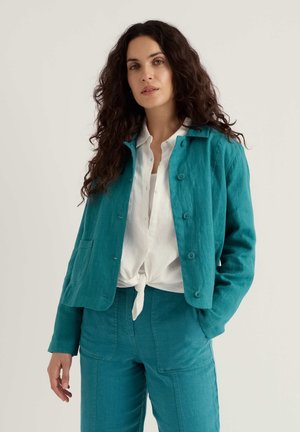 Kvinde med langt krøllet hår iført tealfarvet jakke med knapper og matchende bukser, hvid bluse bundet ved taljen, stående foran en ensfarvet baggrund.