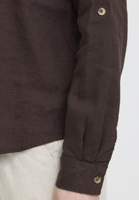 Manchette de chemise marron à manches longues avec détail de bouton portée sur un pantalon beige clair sur un fond uni.