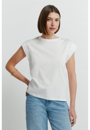 Kobieta z krótkimi brązowymi włosami, ubrana w biały top bez rękawów i jasnoniebieskie dżinsy, stojąca z jedną ręką na biodrze na jednolitym tle.