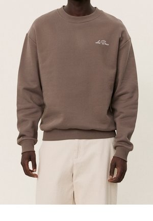 Donkere persoon die een bruine trui met ronde hals met het logo "Les Deux" draagt en licht beige broek, staand tegen een effen achtergrond.