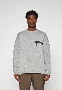 Bărbat cu păr curly scurt, purtând un pulover din fleece gri deschis cu un buzunar cu fermoar negru și pantaloni maronii, stând în fața unui fundal simplu.