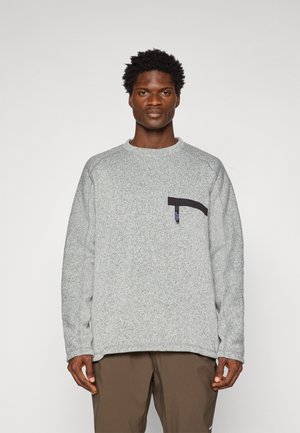 Man with short curly hair wearing a light gray fleece sweater with a black zipper pocket and brown pants, standing against a plain background.