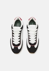 Svart og hvit low-top sneakers med røde detaljer, teksturert semsket skinn og glatt lær. Har hvite snorer og en lys brun gummisåle.