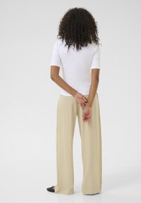 Haut blanc ajusté à manches courtes associé à un pantalon beige à jambes larges. Le modèle se tient debout, les mains jointes derrière le dos, montrant la vue de dos.