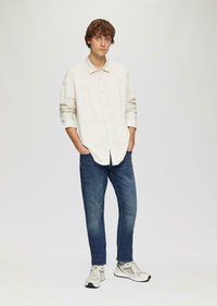 Chemise boutonnée de couleur claire avec les manches retroussées, associée à un jean bleu foncé et des baskets gris clair. Design simple sans motifs visibles.