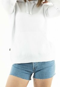 Persona che indossa una felpa bianca con cappuccio e cordini, insieme a pantaloni corti in denim azzurro chiaro con bordi sfrangiati, su uno sfondo bianco.