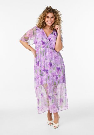 Smilende kvinde med krøllet hår iført en lilla blomsterprint maxi-kjole og hvide åbne hæle, stående med den ene hånd på hoften og den anden nær ansigtet.