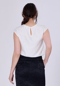 Blouse blanche à manches courtes avec un détail de dos en forme de trou de serrure. Tissu lisse. Portée avec une jupe noire texturée à motifs floraux.