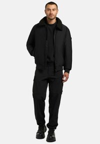 Svart bomberjakke med pelskrage, kombinert med svart genser og cargobukser. Tykt stoff, glidelåslukking og diskrete lommer.