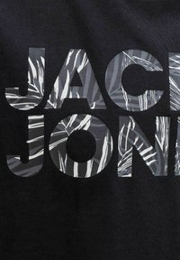 Μαύρο ύφασμα με μεγάλα έντονα γράμματα που σχηματίζουν τη λέξη "JACK JONES", γεμάτα με ένα αφηρημένο μοτίβο φύλλων σε γκρι και λευκό.