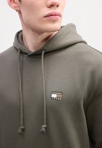 Artimas žmogaus, dėvinčio alyvuogių žalios spalvos sportinę striukę su "Tommy Jeans" logotipo etikete ant krūtinės, vaizdas, matomas kaklas ir apatinė veido dalis.