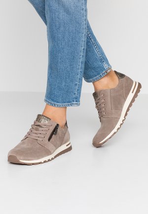Person iført taupe ruskindsneakers med sidelås og hvide gummisåler, kombineret med blå jeans mod en ensfarvet baggrund.
