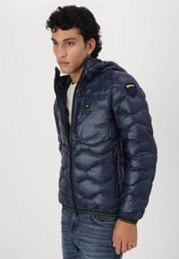 Giacca a vento blu navy con cappuccio, caratterizzata da texture imbottita, zip frontale e accenti gialli a contrasto sulla zip e sul logo.