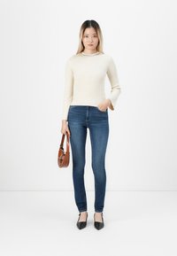Young woman standing against white background wearing cream ribbed sweater, blue skinny jeans, black heels, and holding brown handbag.