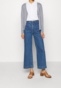 Cárdigan abotonado gris claro sobre una polo blanca, combinado con jeans de mezclilla azul de tiro alto y corte holgado, y sandalias de tacón marrones.