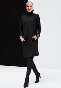 Schwarzes Strickkleid mit hohem Kragen, gerippten Bündchen und seitlichen Reißverschlusstaschen. Das Model kombiniert es mit schwarzen Stiefeln bis zum Oberschenkel.