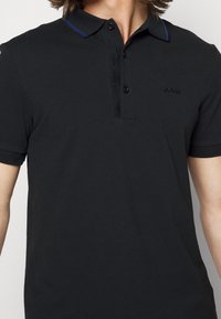 Černé polo tričko vyrobené z texturované tkaniny, s dvouknoflíkovým zapínáním, jemnými modrými akcenty na límečku a vyšívaným logem na hrudi.