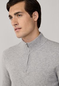 Hombre con cabello corto castaño que lleva un suéter de lana de cremallera gris claro, mirando ligeramente de lado contra un fondo liso.
