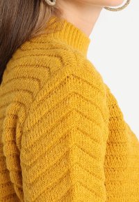 Strickpullover in senfgelb mit hohem Kragen, der ein Zickzackmuster und strukturierten Stoff aufweist. Nahaufnahme des Schulterbereichs.