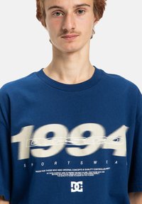 Blå bomulls-T-shirt med djärv vit text som visar året "1994" och ordet "SPORTSWEAR". Klassisk krage och avslappnad passform.