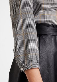 Blouse à carreaux noir et blanc avec des accents orange, dotée d'une manche froncée et d'un détail de bouton, assortie à une jupe en cuir noire.