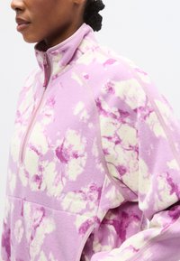 Φλις πουλόβερ σε ροζ χρώμα με λευκό και μωβ σχέδιο σύννεφων, γιακά με μισό φερμουάρ και διακριτικό λογότυπο στο στήθος. Μαλακή υφή.