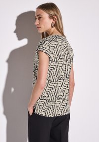 Top a maniche corte color crema con un motivo astratto nero. Presenta un colletto rotondo e una vestibilità comoda, con fessure laterali.