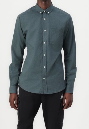 Uomo che indossa una camicia verde scuro a maniche lunghe abbottonata con taschino sul petto e pantaloni neri con tasche laterali.