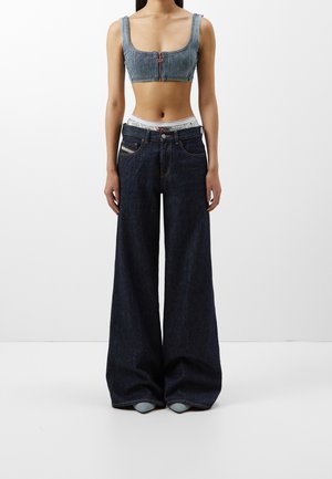 Nainen, jolla on yllään lyhyt harmaa ribbikuvioitu toppi ja ylipitkät tummansiniset leveälahkeiset farkut, seisoo yksinkertaisen valkoisen taustan edessä.