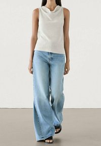 Woman wearing sleeveless light beige top, wide-leg light blue jeans, and black open-toe sandals, standing against a plain background.