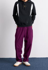 Felpa nera con cappuccio e pannelli laterali bianchi, abbinata a pantaloni larghi viola e sneakers bianche con suole color gomma.