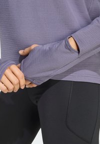 Dugi rukavi ljubičasta majica s rebrastom teksturom i otvorima za palčeve, u kombinaciji s uskim crnim tajicama s kontrastnim šavovima i bočnom džepom.