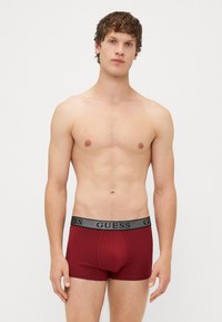 Boxer da uomo rossi in cotone con una cintura elastica grigia che presenta in modo evidente il logo "GUESS" sul davanti.