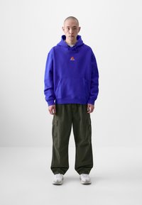 Felpa viola con coulisse e tasca frontale, arricchita da un piccolo logo. Abbinata a pantaloni cargo oliva e sneakers bianche.