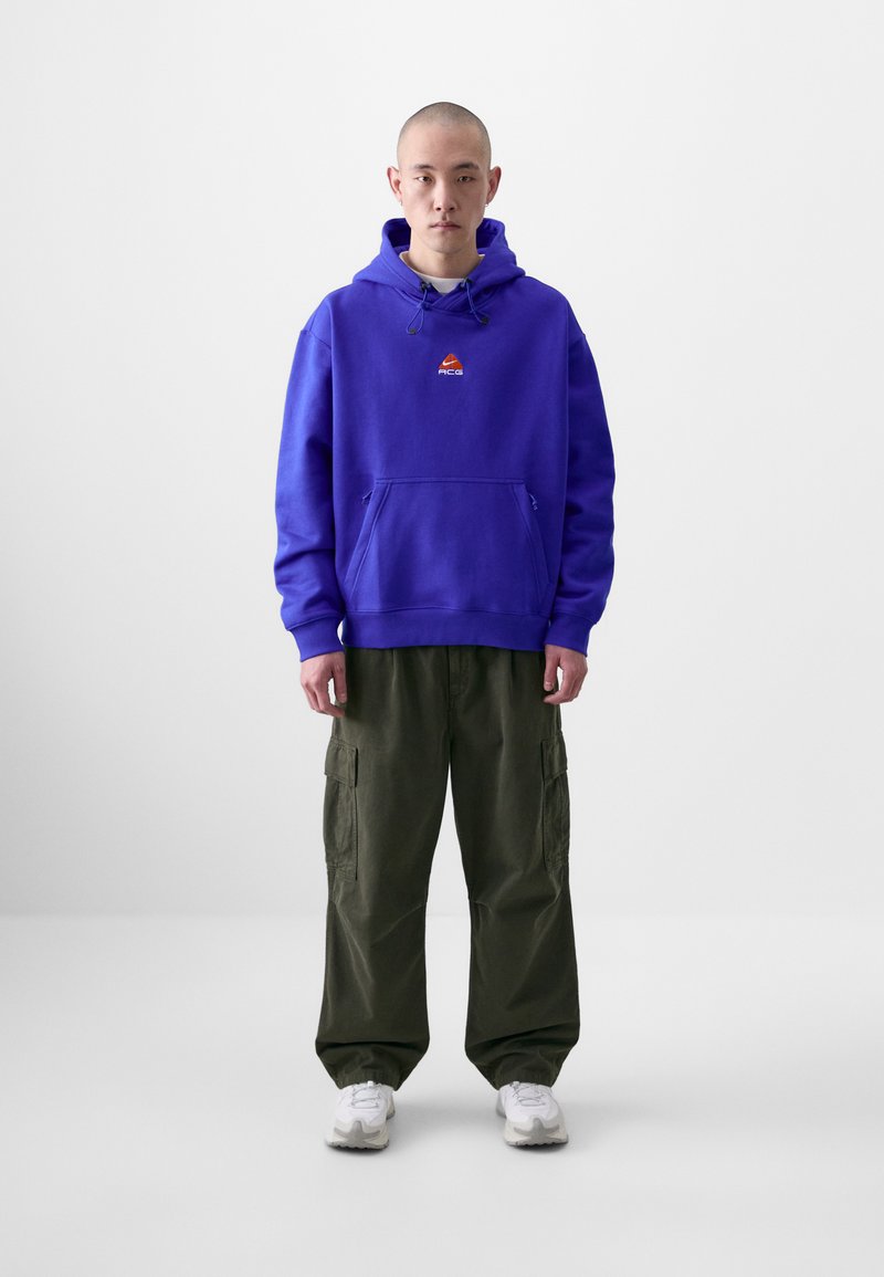 Felpa viola con coulisse e tasca frontale, arricchita da un piccolo logo. Abbinata a pantaloni cargo oliva e sneakers bianche.