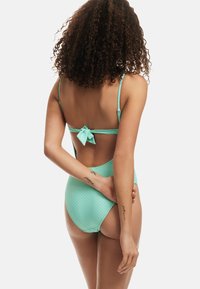 Un bikini taille haute en vert menthe texturé avec des fines bretelles, présentant un détail de nœud à l'arrière. Tissu lisse et design ajusté.