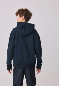 Mørkeblå hættetrøje lavet af blødt stof med stor hætte, ribbede ærmeender og kant. Bagpå er designet enkelt uden mønstre.