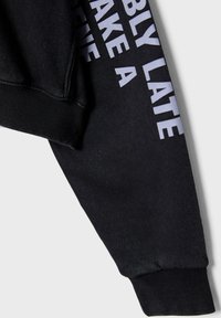 Felpa nera con polsini e orlo a coste, caratterizzata da un audace testo bianco lungo la manica, realizzata in tessuto morbido dalla texture liscia.