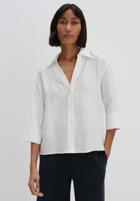 White button-up shirt with a textured fabric, three-quarter sleeves, and a classic collar, paired with dark trousers.