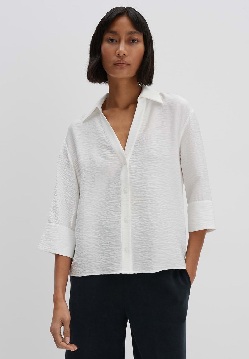 White button-up shirt with a textured fabric, three-quarter sleeves, and a classic collar, paired with dark trousers.