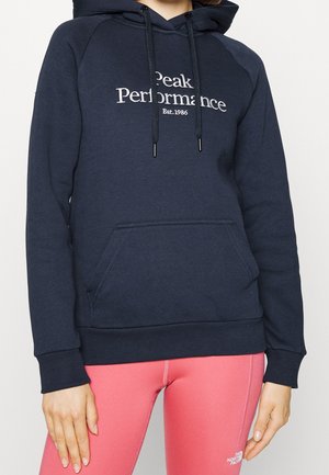 Persoon draagt een navy hoodie met de tekst "Peak Performance Est.1986" en roze legging met het The North Face-logo op de linker dij.