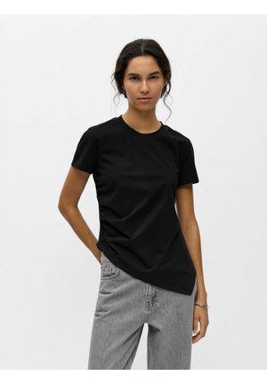 Femme aux cheveux foncés portant un t-shirt noir à manches courtes et un jean gris, debout avec une main dans la poche devant un fond blanc uni.