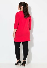 Rote Tunika mit dreiviertelärmel, einfarbig, kombiniert mit schwarzen Leggings und schwarzen High Heels. Glatte Textur, fällt bis zur Mitte des Oberschenkels.