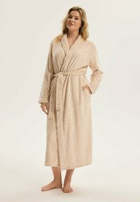 Beżowy, puszysty szlafrok z teksturowanym wzorem, długimi rękawami, paskiem w talii i kieszeniami, sięgający do połowy łydki. Model boso.