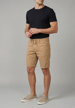 Homme portant une chemise noire à manches courtes, un short beige avec cordon de serrage et des baskets beiges, debout devant un fond gris uni.