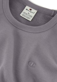 Camiseta de algodón gris con cuello redondo que presenta un pequeño logotipo bordado en la esquina inferior y una etiqueta de cuidados en el interior del cuello.
