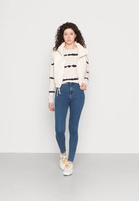 Um colete acolchoado branco sobre uma blusa de mangas longas bege e preta às riscas, combinado com jeans skinny azuis e ténis brancos com detalhes laranja.